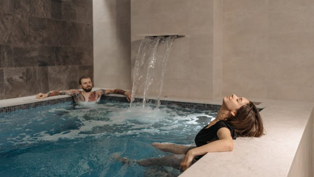 A couple relaxing in an indoor spa pool under a soothing waterfall effect.
