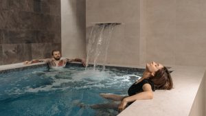 A couple relaxing in an indoor spa pool under a soothing waterfall effect.